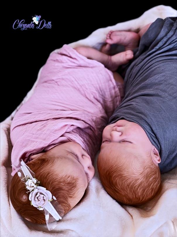 Reborn Max twins cuddled together; The boy and girl both have ginger hair and a fare, rose skin tone. The girl has a rare double crown.