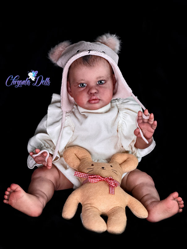 Reborn Ellie full body picture. She is sitting and wearing a pink hat with fuzzy ears. One hand holds a yellow bunny
			and the other hand is waving to camera.